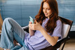 © dikushin - High-angle view of cheerful young woman texting online message using mobile phone sitting on chair in light apartment. Smiling redhead female using cellphone at home on background of window.