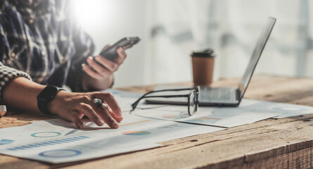 Young accountant hand holding smartphone to call marketing consultants and using laptop computer to analyze sales growth in the global workplace market. accounting concept