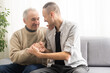 © Angelov - Happy two generations male family old senior mature father and smiling young adult grown son enjoying talking chatting bonding relaxing having friendly positive conversation sit on sofa at home.