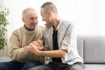 Happy two generations male family old senior mature father and smiling young adult grown son enjoying talking chatting bonding relaxing having friendly positive conversation sit on sofa at home.