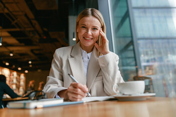 Wall Mural - Smiling businesswoman drinking coffee and making notes while working in cafe