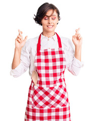 Poster - Beautiful young woman with short hair wearing professional cook apron gesturing finger crossed smiling with hope and eyes closed. luck and superstitious concept.