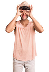 Poster - Young hispanic man wearing summer hat doing ok gesture like binoculars sticking tongue out, eyes looking through fingers. crazy expression.