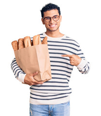 Hispanic handsome young man holding paper bag with bread smiling happy pointing with hand and finger