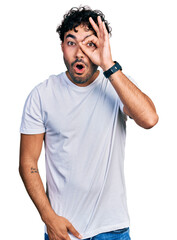 Poster - Hispanic young man with beard wearing casual white t shirt doing ok gesture shocked with surprised face, eye looking through fingers. unbelieving expression.