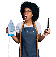 Canvas Print - Young african american woman dressmaker designer wearing atelier apron holding iron and scissors angry and mad screaming frustrated and furious, shouting with anger. rage and aggressive concept.