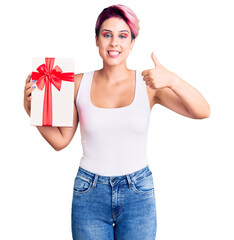 Sticker - Young beautiful woman with pink hair holding gift smiling happy and positive, thumb up doing excellent and approval sign