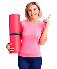 Poster - Young blonde woman holding yoga mat surprised with an idea or question pointing finger with happy face, number one