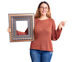 Sticker - Beautiful young woman holding empty frame smiling happy pointing with hand and finger to the side