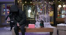 Person In Parka With Hood Pulled Low, Hiding Face, Wearing Sunglasses. Suspicious Guy Brings Box In Wrapping Paper And Leaves It On Bench Next To Dachshund Dog In Knitted Sweater
