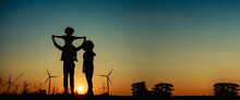 Silhouettes Of Happy Family Father, Mother And Child Daughter Sits On The Shoulders Of His Father With Windmills For Electricity Generation At Sunrise.