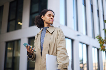 Confident and stylish young African American pretty woman in casual clothes, looking away, staying on the city street, holding modern mobile phone in hands