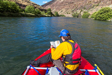 A Man Dressed In A Yellow Dry Suit Reads A Map On The Front Of An Oar Raft On The Deschutes River In Oregon On A Sunny Day.