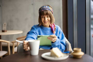 Wall Mural - Young stylish woman works on digital tablet while sitting relaxed with coffee and cake at coffee shop. Concept of remote creative work from public place