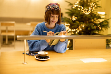 Wall Mural - Young adult woman works on a digital tablet while sitting at modern coffee shop in yellow tones with Christmas tree on background. Concept of remote creative work online