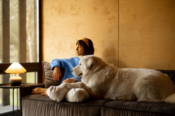 Canvas Print - Woman cares her dog while sitting on a couch by the window, spending leisure time together in wooden house on nature