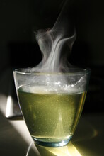 Transparent Glass Containing Green Hot Herbal Tea Hit By A Sun Ray. Steam Motion On Dark Background.