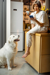 Young woman uses smartphone and drinks a coffee while sitting with her dog on kitchen at home. Concept of morning routine and domestic lifestyle