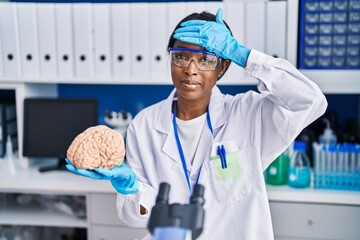 Poster - African young woman working at scientist laboratory holding brain stressed and frustrated with hand on head, surprised and angry face