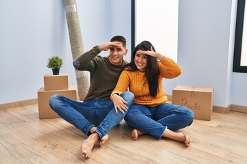 Poster - Young couple sitting on the floor at new home very happy and smiling looking far away with hand over head. searching concept.