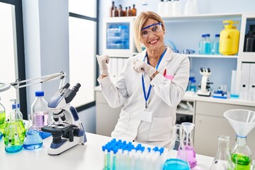 Wall Mural - Middle age blonde woman working at scientist laboratory pointing to the back behind with hand and thumbs up, smiling confident