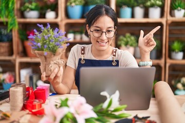 Canvas Print - Young hispanic woman working at florist shop doing video call cheerful with a smile on face pointing with hand and finger up to the side with happy and natural expression