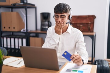 Wall Mural - Young hispanic man working using computer laptop holding credit card laughing and embarrassed giggle covering mouth with hands, gossip and scandal concept