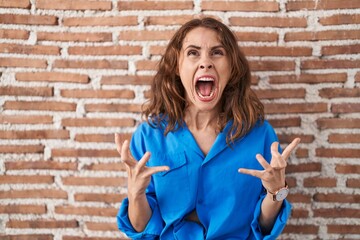 Sticker - Beautiful brunette woman standing over bricks wall crazy and mad shouting and yelling with aggressive expression and arms raised. frustration concept.