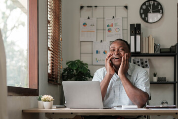 Wall Mural - Smiling black man using laptop at home in living room. Happy mature businessman send email and working at home. African american freelancer typing on computer with paperworks and documents on table.