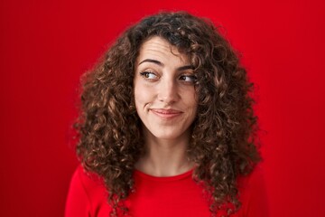 Sticker - Hispanic woman with curly hair standing over red background smiling looking to the side and staring away thinking.