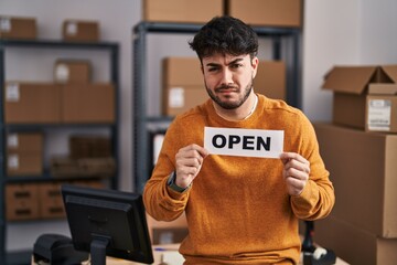 Canvas Print - Hispanic man with beard working at small business ecommerce holding open sign clueless and confused expression. doubt concept.