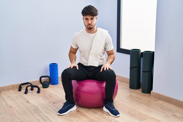 Wall Mural - Hispanic man with beard sitting on pilate balls at yoga room puffing cheeks with funny face. mouth inflated with air, crazy expression.