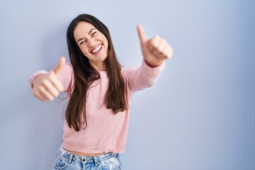 Sticker - Young brunette woman standing over blue background approving doing positive gesture with hand, thumbs up smiling and happy for success. winner gesture.