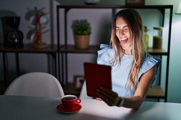 Canvas Print - Young hispanic woman using touchpad sitting on the table at night sticking tongue out happy with funny expression. emotion concept.