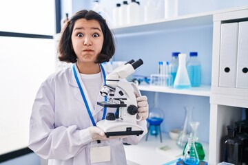 Sticker - Young hispanic woman working at scientist laboratory holding microscope puffing cheeks with funny face. mouth inflated with air, catching air.