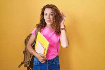 Sticker - Young caucasian woman wearing student backpack and holding books smiling with hand over ear listening an hearing to rumor or gossip. deafness concept.