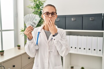 Poster - Young doctor woman holding dollars banknotes covering mouth with hand, shocked and afraid for mistake. surprised expression