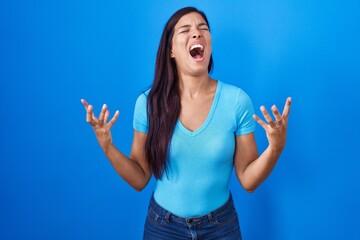 Wall Mural - Young hispanic woman standing over blue background crazy and mad shouting and yelling with aggressive expression and arms raised. frustration concept.