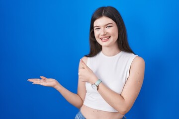 Wall Mural - Young caucasian woman standing over blue background showing palm hand and doing ok gesture with thumbs up, smiling happy and cheerful