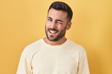 Wall Mural - Handsome hispanic man standing over yellow background winking looking at the camera with sexy expression, cheerful and happy face.