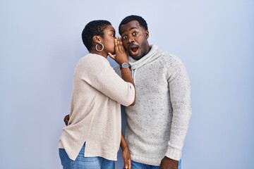 Young african american couple standing over blue background together hand on mouth telling secret rumor, whispering malicious talk conversation