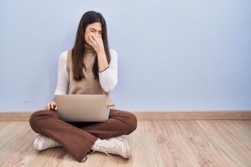 Poster - Young brunette woman working using computer laptop sitting on the floor smelling something stinky and disgusting, intolerable smell, holding breath with fingers on nose. bad smell