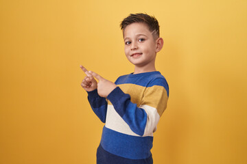 Little hispanic boy standing over yellow background smiling and looking at the camera pointing with two hands and fingers to the side.