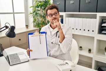 Sticker - Young hispanic man wearing doctor stethoscope holding clipboard serious face thinking about question with hand on chin, thoughtful about confusing idea