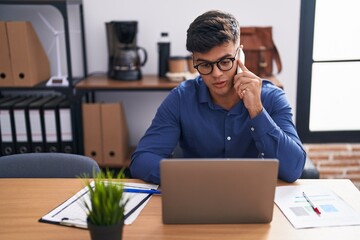 Wall Mural - Young hispanic man business worker using laptop talking on smartphone at office