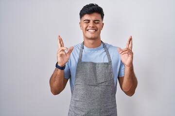 Poster - Hispanic young man wearing apron over white background gesturing finger crossed smiling with hope and eyes closed. luck and superstitious concept.