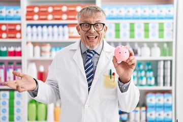 Sticker - Hispanic man with grey hair working at pharmacy drugstore holding piggy bank celebrating achievement with happy smile and winner expression with raised hand