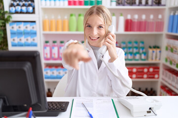 Sticker - Young caucasian woman working at pharmacy drugstore speaking on the telephone pointing to you and the camera with fingers, smiling positive and cheerful