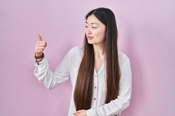 Wall Mural - Chinese young woman standing over pink background looking proud, smiling doing thumbs up gesture to the side