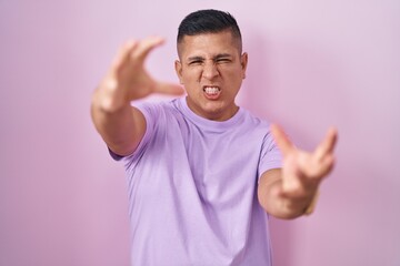 Sticker - Young hispanic man standing over pink background shouting frustrated with rage, hands trying to strangle, yelling mad